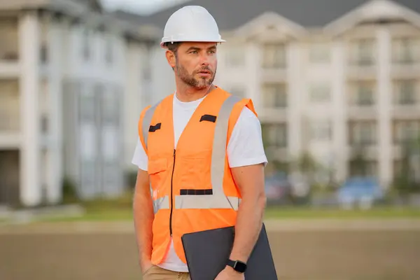 Engineer with clipboard, building inspection. Builder at building site. Construction manager or engineer in helmet outdoor. Male construction engineer. Architect or engineer at a construction site