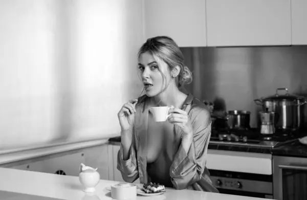 Sexy woman sitting in the kitchen in the morning and drinking coffee. Sensual housewife with bare breast lick spoon