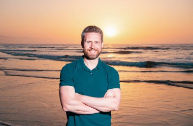 Gün batımında sahilde mutlu bir adamın portresi. Seyahat tatili konsepti. Yaz tatili. Sahilde yaz tatilinin tadını çıkaran neşeli gülümseyen adamın portresi.