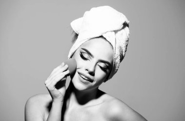 Yüzünde sünger olan bir kadın. Yanağa, hatlara ve fosforlu kalemlere toner uygula. Yüz kremi ürünü, tonal fondöten. Kafasında havlu, yüzünde parlak makyaj olan bir model.