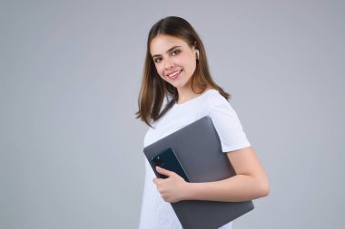 Mutlu Öğrenci. Beyaz tişörtlü, dizüstü bilgisayarı ve bilgisayarı olan genç bir kız, gri arka planda izole edilmiş. Stüdyoda poz veren öğrenci. Video görüşmesi yapan öğrencinin portresi. Öğrencilerin zamanı.