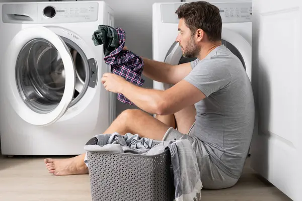 Man with clothes near washing machine. Handsome man sits in front of washing machine. Loads washer with dirty laundry. Man cleaning clothes. Housework for single alone guy. Home laundry