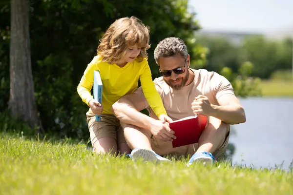 Father and son sitting on green grass in garden and reading book together. Happy family reading book together in green nature. Father is sitting with son read book. Kid is very interested