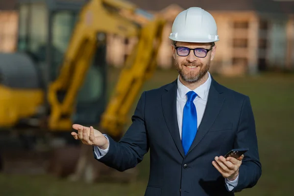 Civil engineer worker at a construction site. Mature engineer worker. Man in suit and hardhat helmet at construction site. Middle aged head civil engineer worker standing outside near excavator