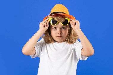 Mavi stüdyoda, şapkalı, tişörtlü ve güneş gözlüklü bir çocuğun moda portresi.