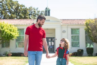 İlkokulun ebeveynleri ve öğrencileri el ele gider. Tişörtlü öğretmen ve okul parkının yanında sırt çantalı sevimli okul çocuğu.