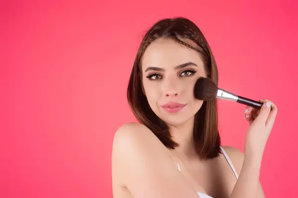 Girl hold blush blusher apply powder visage isolated over studio background. Young woman powdering cheeks. Makeup brush. Female model gets blush powder on the cheekbones