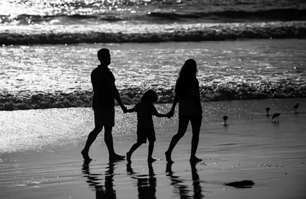 Silhouette of family walking on beach. Parents and kid walking on golden sand, mom and dad holding girls hands. Full length. Family outdoor activities concep