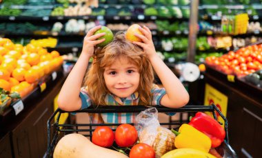 Çocuk dükkanda elma seçiyor. Komik küçük çocuk gülüşü ve elma tutuşu. Çocuk süpermarkette alışveriş yapıyor. Arabalı küçük çocuk marketten taze sebze seçiyor.