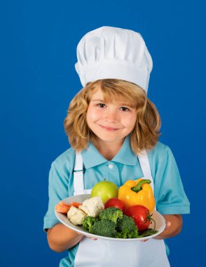 Aşçı şapkalı ve önlüklü çocuk yemek hazırlayan sebzeli yemek tabağı. Mutfakta sebzeli küçük bir aşçı. Doğal çocuk yemekleri.