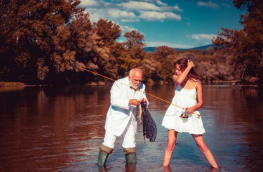 Genç seksi kadın ve yaşlı balıkçı elinde oltayla nehirde duruyor. Baba ve kızı balık tutuyor. Olgun zengin balıkçı takım elbiseli olta kamışlı, nehirde dönen makaralı.