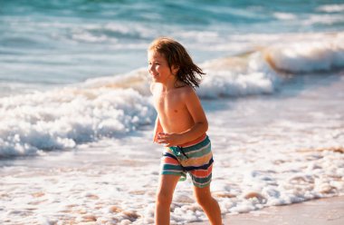 Kumsalda koşan küçük çocuk mavi denizde su sıçratıyor. Çocuk yaz plajında yürüyor.