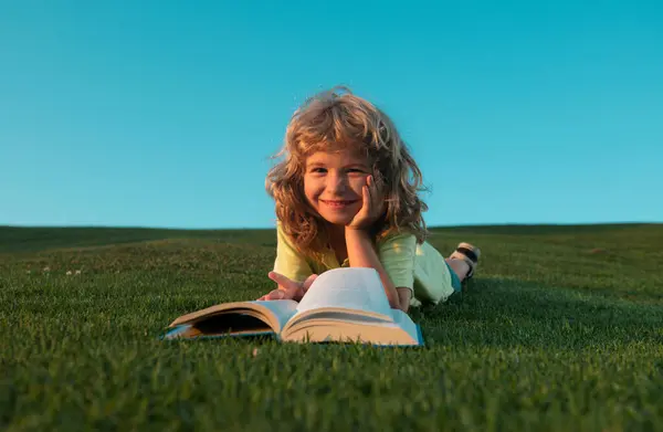 Smart kid boy reading book in park outdoor. Smiling funny child in t-shirt having fun reading book in park