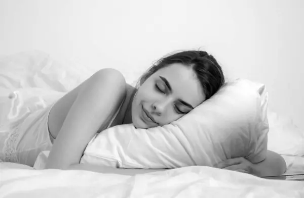 Young woman sleeping well in bed hugging soft white pillow. Girl resting, good night sleep. Woman sleeping. Beautiful young woman lying in bed and keeping eyes closed while covered with blanket