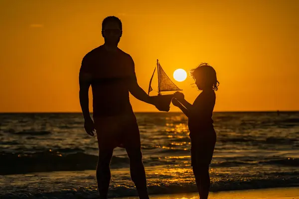 Father and son playing with toy sailboat, toy ship yacht on sea beach at summer sunrise. Sunset silhouette of father and son dreaming on cruise. Dreaming to travel with kid son. Fathers day