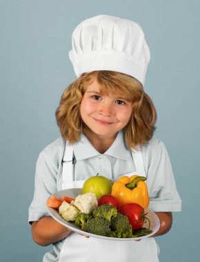 Çocuk aşçı, sebzeli tabak hazırlar. Gri stüdyo arka planında yemek hazırlar. Çocuklar yemek yapıyor. Önlüklü ve şapkalı genç çocuk mutfakta sağlıklı bir sebze yemeği hazırlıyor.