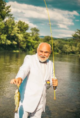 Portrait of cheerful senior man fishing. Grandfather with catch fish. Mature man fisherman in white suit and bowtie with fishing rod, spinning reel on river. Catching trout fish