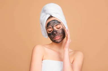 Kömür maskeli bir kadın burnuna dokunuyor. Çamur maskesi, yüz maskesi kaplıcası. Kozmetik çamur yüz prosedürü olan güzel bir kadın, spa sağlık konsepti. Cilt bakımı güzellik tedavisi. Havlu kafaya.