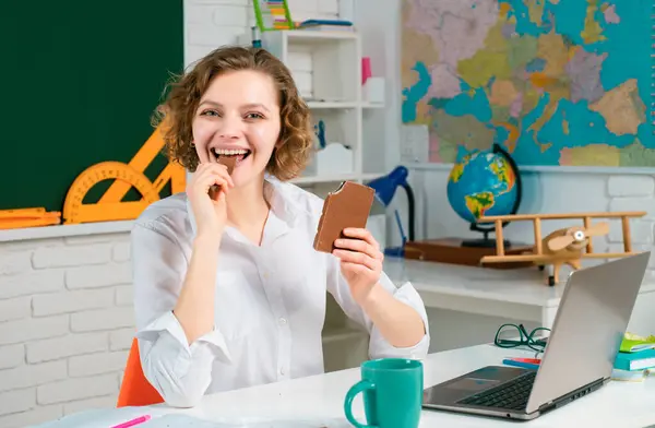 Portrait of smiling female teacher eat chocolate near blackboard in classroom at school