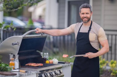 Arka bahçede ızgarada barbekü yapan yakışıklı bir adam. Açık hava yemekleri ve Amerikan yaşam tarzı konsepti. Yakışıklı adam barbekü eti hazırlıyor. Dışarıda yemek ve yemek pişirme kavramı