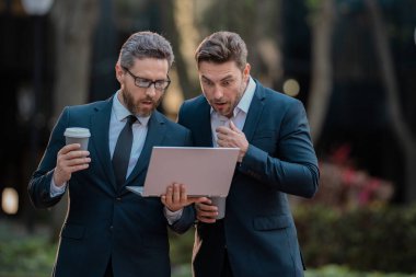 Two surprised businessman using laptop outdoor. Handsome business man in suits working on laptop and communication with business projects in city. Partners thinking and plan for business clipart