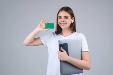 Yüzü ve dizüstü bilgisayarının yanında kredi kartı tutan beyaz tişörtlü genç bir kadın. Gri arka planda izole edilmiş. Zeka ya da banka kartını öde. Banka kartı. Plastik kredi kartı