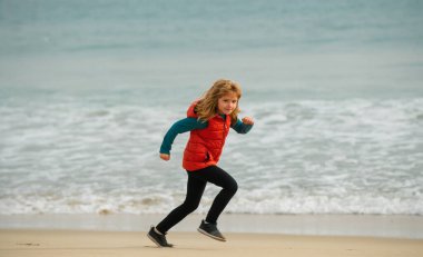 Sahilde oynayan çocuklar. Çocuklar yaz tatilinde denizde oynarlar. Kum ve su eğlencesi, güneş koruması. Okyanus kıyısında koşan ve zıplayan küçük bir çocuk. Çocuklarla sabah koşusu