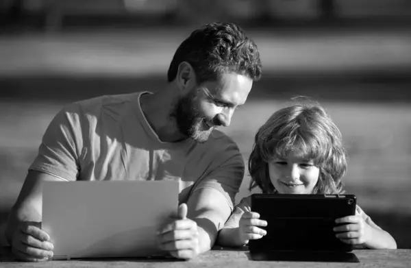 Father teaching son to use laptop, dad and school boy child looking computer screen and tablet, playing game, watching video, sitting on grass. Outdoor family weekend