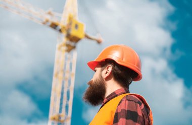 Male construction worker in work clothes and a construction helmet. Improvement and renovation. Brutal man builder. Engineer builder in uniform. Construction crane