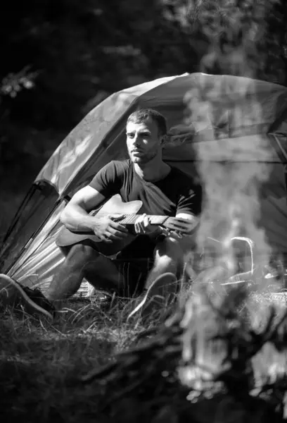 Romantic guy camping outdoors and sitting near tent. Handsome man playing guitar in forest with bonfire. Country music song. Guy by a fire in the forest, weekend near campfire
