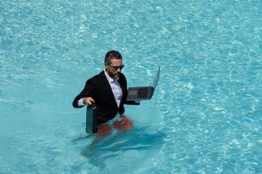 Deniz sahilinde bilgisayarla çalışan takım elbiseli komik bir işadamı. Yaz tatili, açık hava ve iş seyahati konsepti. Çılgın ofis çalışanı. Dizüstü bilgisayarda çalışan bir adam havuz kenarında kumaş içinde oturuyor.