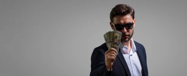 İyi ödeme. Yakışıklı adam gri stüdyoda izole edilmiş para tutuyor. Dolar banknotları halinde para. Yığınla para, finans, yatırım ve tasarruf. Model pankartı