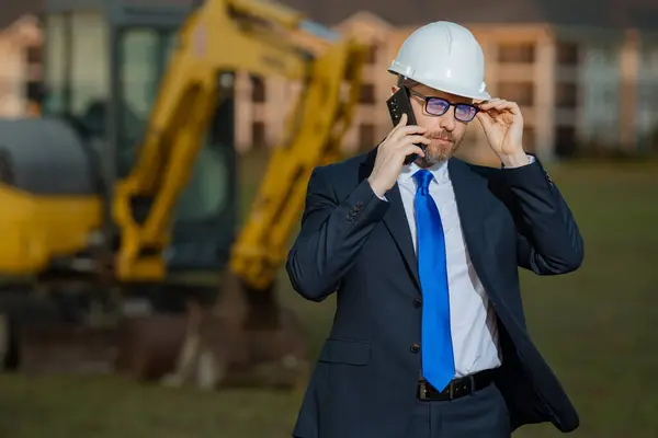 Successful construction business owner. Construction worker in suit and helmet near excavator. Confident construction owner in front of construction site. Civil engineer worker
