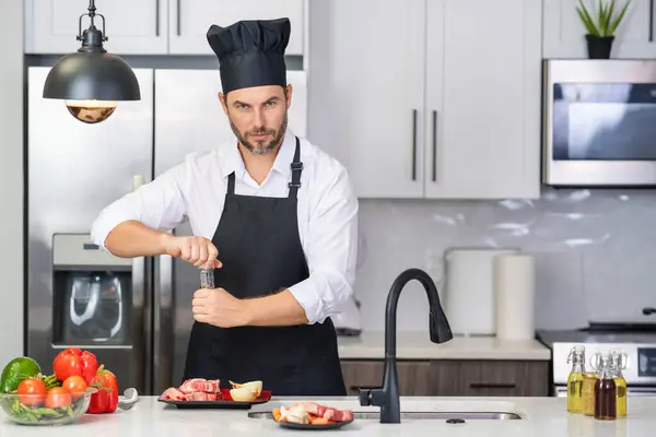キッチンでハンサムな男は健康的なフルーツサラダと肉を準備しています エプロンの男とシェフの帽子はキッチンで食べ物を準備しています シェフの帽子のシェフは キッチンでおいしい料理を調理します モダンなキッチンインテリア — ストック写真