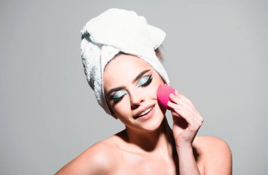 Yüzünde sünger olan bir kadın. Yanağa, hatlara ve fosforlu kalemlere toner uygula. Yüz kremi ürünü, tonal fondöten. Kafasında havlu, yüzünde parlak makyaj olan bir model.