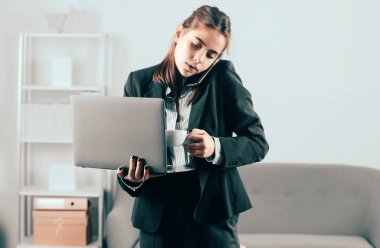 İş kadını ofiste dizüstü bilgisayarını kullanarak oturuyor. Ofis çalışanı kahve içiyor, telefonla konuşuyor, dizüstü bilgisayar kullanıyor.