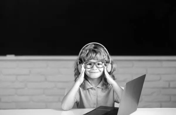 ヘッドフォンの賢い男の子は机に座って 学校でラップトップでオンラインで勉強し 賢い子供はインターネットのレッスンを使ってノートパソコンでイヤホンを身に着けています — ストック写真