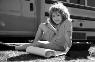 Okul otobüsünün yanında elinde tabletle çimlere uzanan mutlu okul çocuğu.