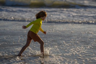 Çocuk yaz günü tropikal plajda koşuyor ve oynuyor. Çocuk açık hava aktivitelerinin tadını çıkarıyor. Denizde yaz tatili. Gün batımında sahilde koşan küçük bir çocuk. Yaz tatilindeki çocuklar