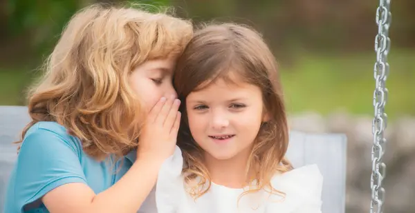 Little boy whispers to lovely girl in ear. Two children outdoors. Portrait of adorable brother and sister smile and laugh together. Happy lifestyle kids