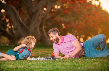Baba oğul çim parkında çimlerde satranç oynuyorlar. Babalar Günü, aileyi sevmek, ebeveynlik, çocukluk konsepti. Akıllı çocuk, akıllı çocuklar.