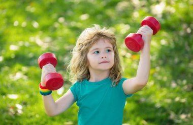 Dambıl kaldıran çocuk. Dambılı şirin çocuk eğitimi. Çocuk fitness. Çocuk dışarıda halterlerle egzersiz yapıyor. Komik heyecanlı çocuk sporcusu