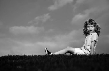 Çimlerin üzerinde oturan şirin bir yüz ifadesi olan küçük bir çocuk. Neşeli çocuk yeşil yaz çayırında eğleniyor.