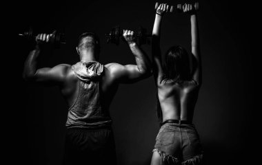 Kaba bir çift halterleriyle birlikte egzersiz yapıyor. Seksi, formda bir vücut. Çıplak vücutlu kaslı bir adam, arka planda koyu renkli halterleri olan fitness kadını. Dambılı çift eğitimi