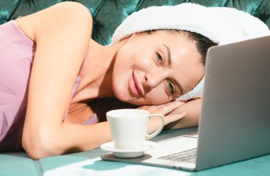 Kahveli ve dizüstü bilgisayarlı güzel ve şehvetli genç bir kadın pijamalı. Yatak odasında boş zamanlarının tadını çıkarıyor.