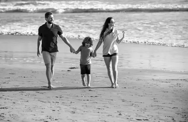 Full length of family of three walking along the sea shore. Parents holding hands of son and walking on beach