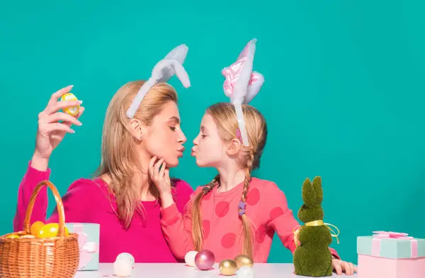 Happy easter. A mother and her daughter painting Easter eggs and kissing. Happy family preparing for Easter. Cute little child girl wearing bunny ears on Easter day isolated on blue