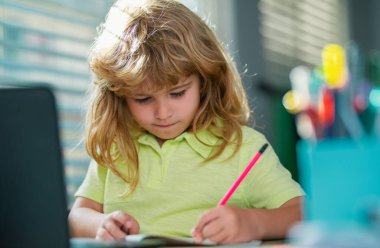 Serious school child writing homework in school class. Portrait of school kid boy siting on table doing homework. Child holding pencil and writing. Boy drawing on white paper at the table