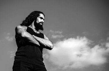 Young muscular handsome man standing with arms folded against blue sky looking forward copy space and space for text. Fashionable man with long wavy hair and strong healthy body