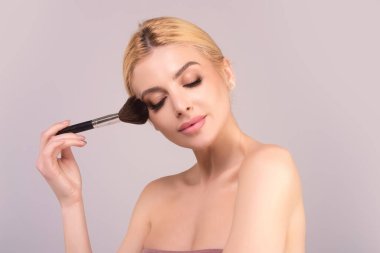 Studio portrait of a woman applying cosmetic tonal foundation on face using makeup brush. Beautiful girl doing contouring apply blush on cheeks isolated on studio background clipart